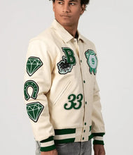 Load image into Gallery viewer, Man wearing a cream varsity jacket with green letterman patches on a gray background
