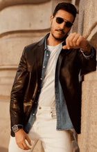 Load image into Gallery viewer, Man wearing sunglasses, leather jacket, denim shirt, and white pants against a textured wall.
